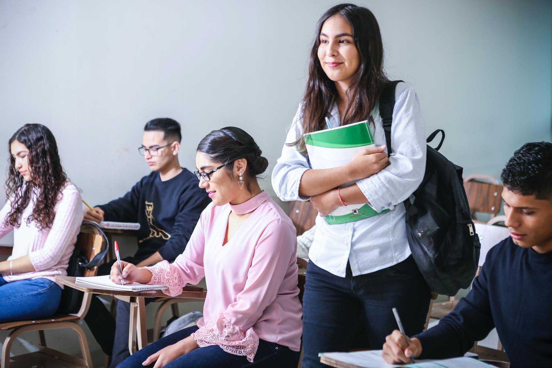 Students in classroom, writing at desks, girl looking around for a seat