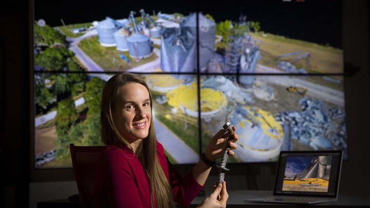 Christine Wittich holds a damaged anchor bolt in front of a photo of damaged steel grain bins.