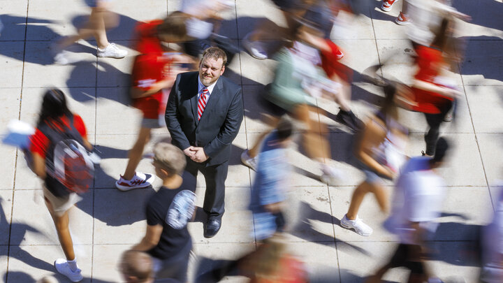 A man in a black suit with a red tie stands in the middle of a blurry people walking around him.