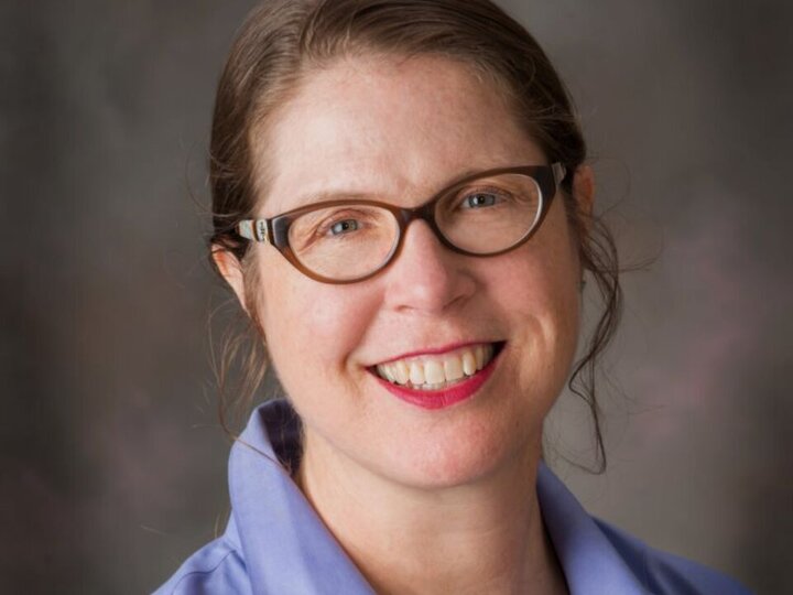 A woman smiles at the camera. She is wearing brown glasses, a blue collared shirt, and her hair is pulled back in a low ponytail.