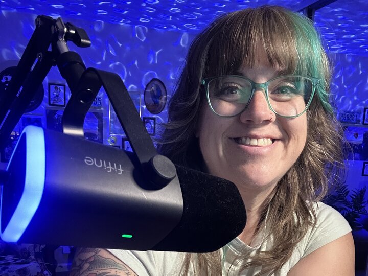 Smiling person with glasses near a microphone, surrounded by blue ambient lighting and wall decor.