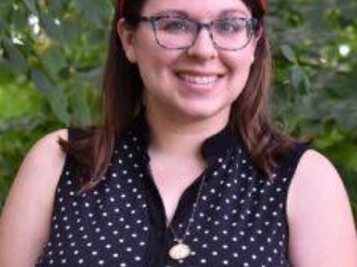 Woman wearing glasses, a red headband, and a black polka-dotted top, smiling against greenery.